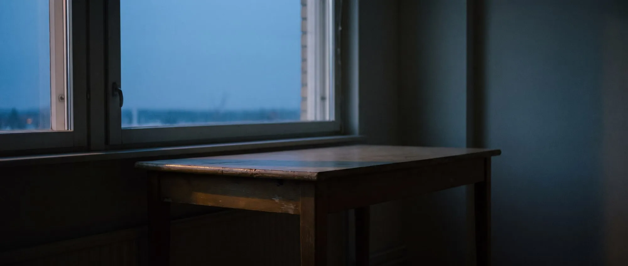 A window at dusk from inside a dim room, the last light catching the edge of a wooden table.
